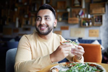 Young smiling man with slice of pizza sitting by table in cafe and enjoying lunch time