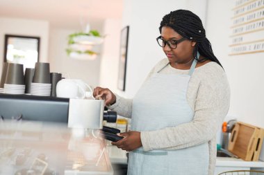 Kahve makinesinin yanında duran ve kafeterya müşterileri için sıcak aromatik içecek hazırlayan iş elbiseli genç bir kadın.