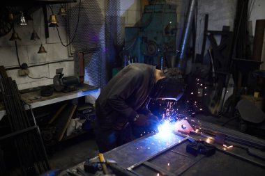 Maskeli meşgul işçi, karanlık atölyede çerçeve içinde katılırken metal plakalarla çalışan kaynak prosesine yoğunlaştı