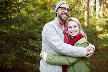 Portrait of young attractive loving Caucasian couple standing in conifer forest,  hugging each other and smiling at camera happily clipart