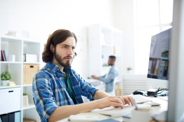 İki masaüstü bilgisayarlarla çalışma ve Office masada otururken klavyede yazarak bilgisayar programcısı ciddi konsantre hipster genç sakallı