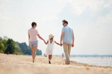 Yaz tatili sırasında sahilde yürüyüş yaparken el ele tutuşan mutlu ailenin geri görünümü