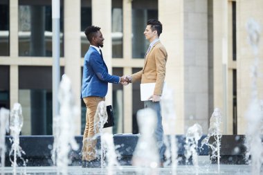 Başarılı anlaşma ile birbirlerini tebrik ederken el sıkışma yapan pozitif başarılı işadamları, ofis binası meydanının yanında duruyorlar.