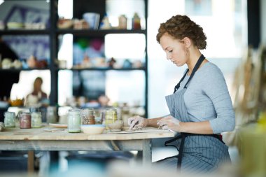 Çalışma odasındaki genç yaratıcı kadın kil zanaat atölyesinde masa başında oturuyor ve toprak eşyalar için küçük detaylar hazırlıyor.