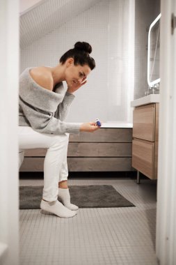 Upset shocked attractive young woman with hair bun sitting on toilet bowl in bathroom and looking at pregnancy test, she not believing her eyes clipart