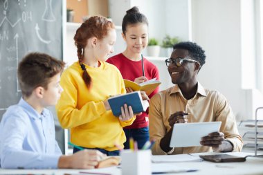 Yeni kelimeleri tartışırken tabletle öğretmenlerinin yanında duran kitap veya sözlük ve kalemleri olan öğrenci grubu
