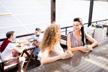 Açık hava kafede bar tezgahı tarafından duran iki dost kız, içki içiyor ve arka planda erkeklerle haber tartışıyor
