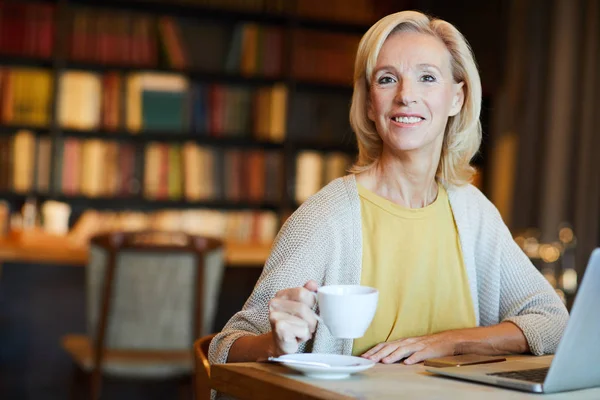 Happy blonde woman with cup of tea or coffee sitting in front of laptop in cafeteria