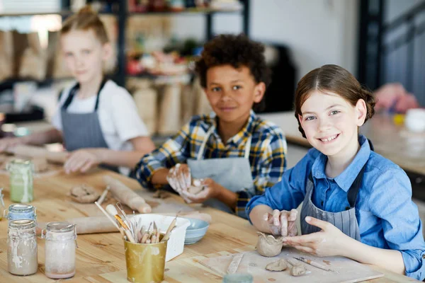 テーブルのそばに座って 背景に彼女の学校の同級生と粘土カップを作っている間 幸せな小さな女の子があなたを見て — ストック写真