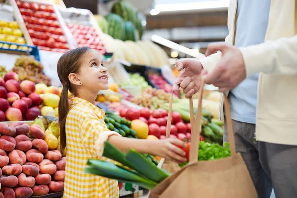 黄色いドレスを着た陽気なかわいい女の子は 野菜を袋に入れ 農家市場で買い物を手伝いながら父親を見ている — ストック写真