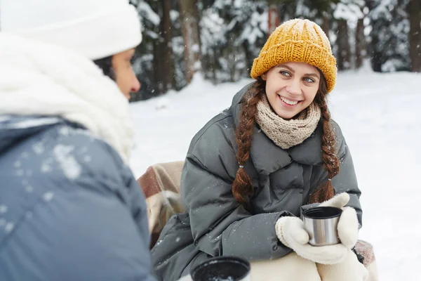 黄色いニット帽をかぶったポジティブな可愛い女の子が 冬の森でキャンプでサーモスマグからお茶を飲みながら友達に話しかける — ストック写真
