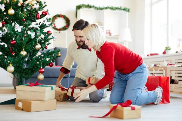 一对年轻夫妇在圣诞前夕把大大小小的礼品盒放在装饰过的圣诞树下 — 图库照片