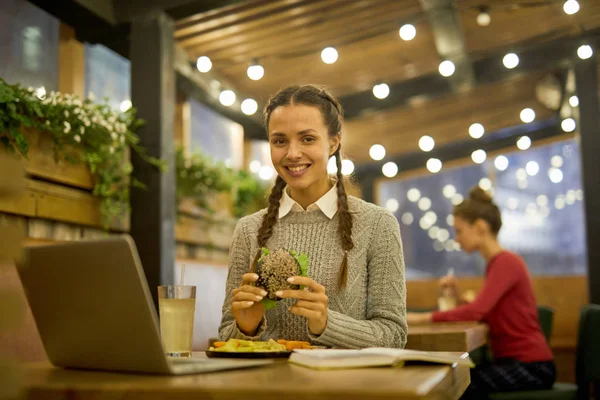 Усміхнений Студент Гамбургером Сидить Столом Перед Ноутбуком Обідає Готується Семінару — стокове фото