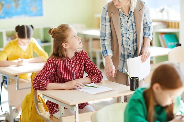 木製の机に座って 教室でのテスト中に彼女に助言しながら 教師を見てチェッカーシャツの小学生の女の子 — ストック写真