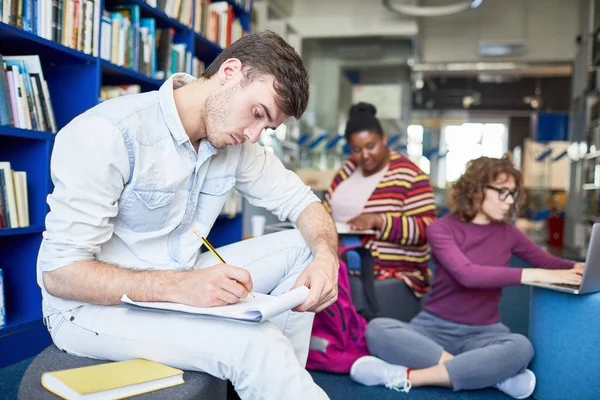 Skoncentrowany Mężczyzna Student Piśmie Raport Siedząc Bibliotece Innymi Wieloetnicznej Studentów — Zdjęcie stockowe