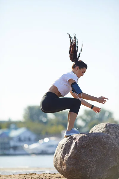 Тренировка Пляже Привлекательная Атлетичная Молодая Женщина Повязкой Смартфоне Прыгающая Камне — стоковое фото