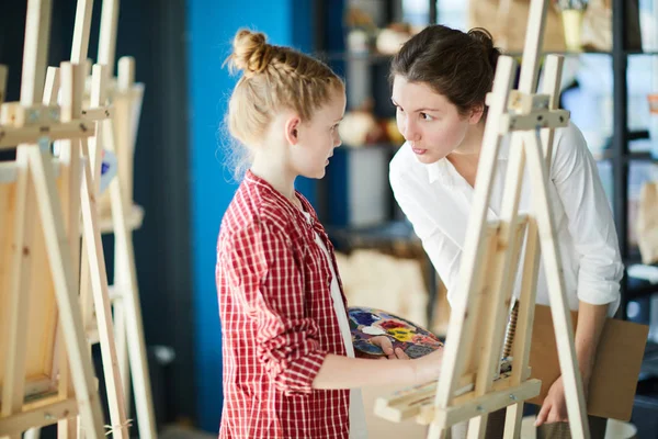 絵画のレッスンでイーゼルのそばに立っている間 彼女の先生に耳を傾けるカラーパレットを持つ小さな女の子 — ストック写真