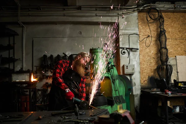 严重集中英俊的胡须男子在围裙使用研磨机 同时切割金属细节在办公桌上用各种手工工具 火花喷泉吹出 — 图库照片