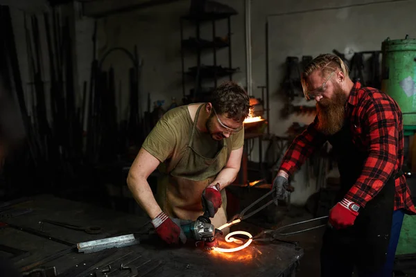 两名专业铁匠穿着工作服站在铁锤前 在铁匠中加工热熨斗工件 — 图库照片