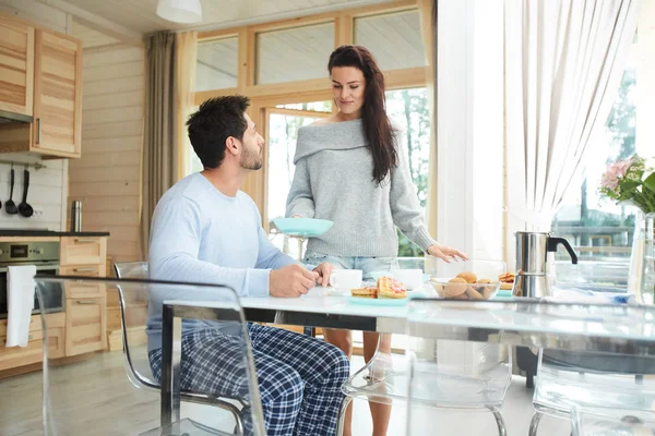 夫に朝食を運び テーブルの上にプレートを置く黒髪の慎重な魅力的な若い妻 キッチンで彼の朝食を待っているハンサムな夫 — ストック写真