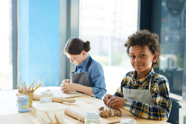 Two intercultural kids in casualwear sitting by desk, kneading clay and making creative things at lesson of crafts