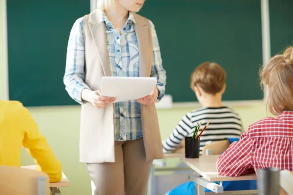 年轻教师一边在一排学生的课桌之间来回走动 一边拿着文件 同时为班里的小学生布置任务 — 图库照片