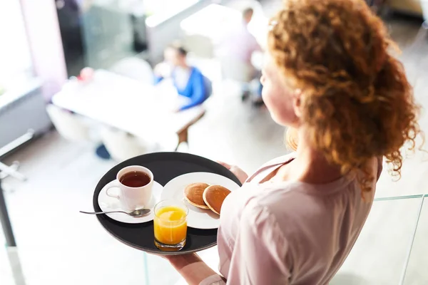 パンケーキとお茶とジュースを運んでいる若い女性がカフェのテーブルの一つに移動しながら — ストック写真