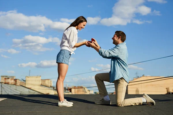 幸せな愛情の男は1膝に乗って 都市の屋根の上に美しい女性にプロポーズをしながら婚約指輪とオープニングジュエリーボックス — ストック写真