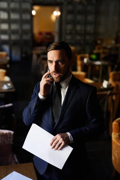 彼とプロジェクトについて話し合いながら 正式なスーツを着た若いあごひげのビジネスマンが電話で同僚の話を聞き テーブルに立って書類を持っている — ストック写真