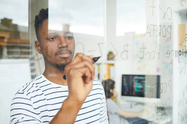 Serious concentrated handsome young African-American web developer using programming language for coding and writing it on glassy wall in office