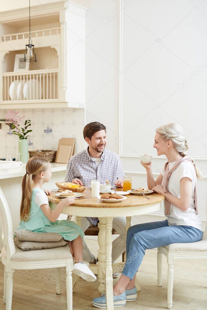 Cheerful talkative young family in casual clothing enjoying breakfast and communication while sitting at table sharing stories in cozy kitchen