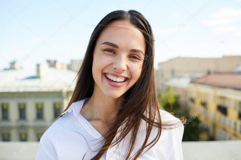Cheerful excited pretty lady with hair blown by wind laughing and looking at camera while standing on roof in city