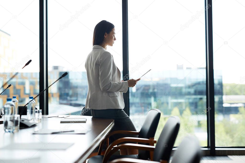 Side view portrait of elegant businesswoman using digital tablet standing alone in office and leaning on meeting table against window, copy space