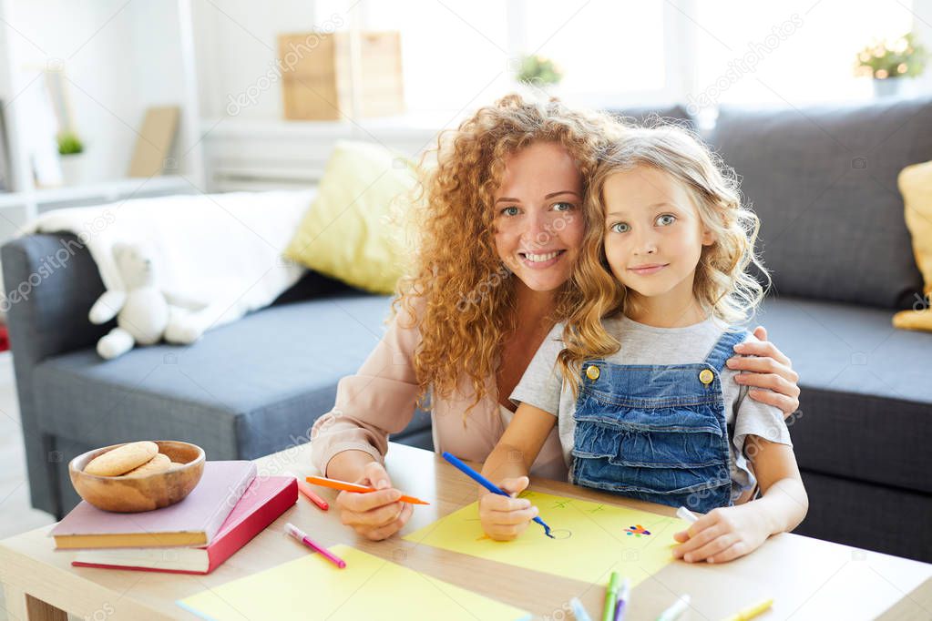 Young cheerful woman and her daughter sitting by table and drawing with highlighters in living-room