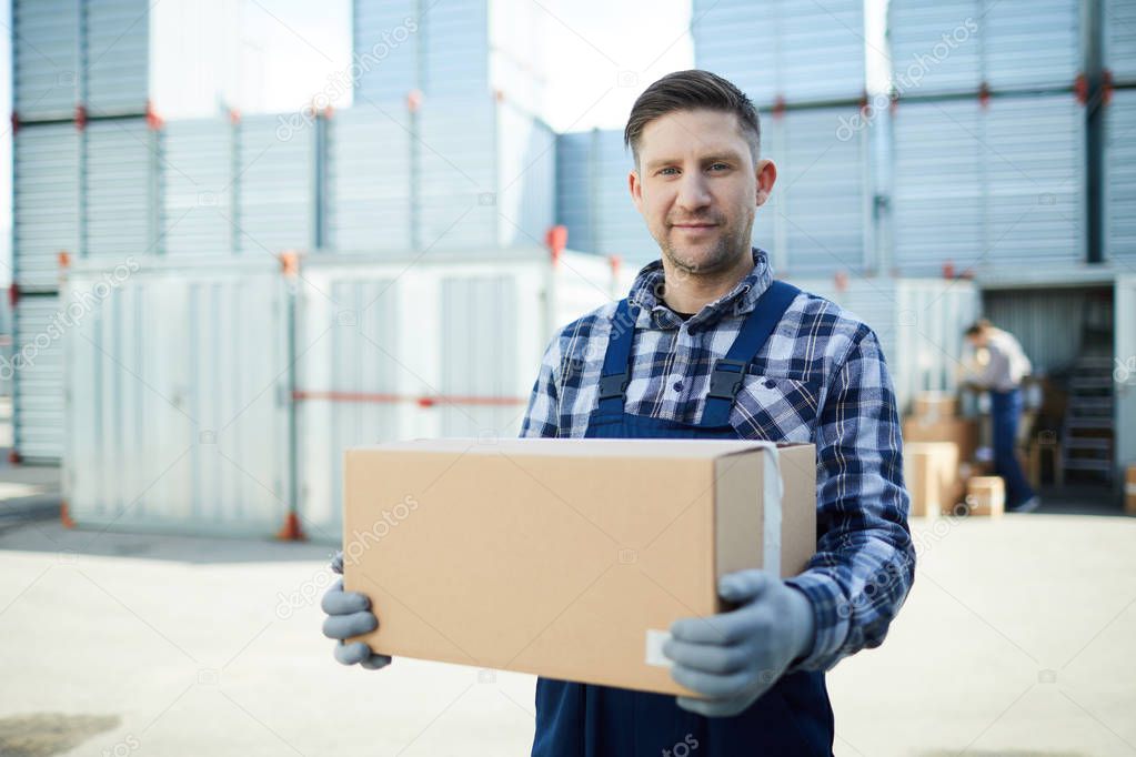 Content handsome manual worker in workwear holding cardboard box and looking at camera while working at container storage area