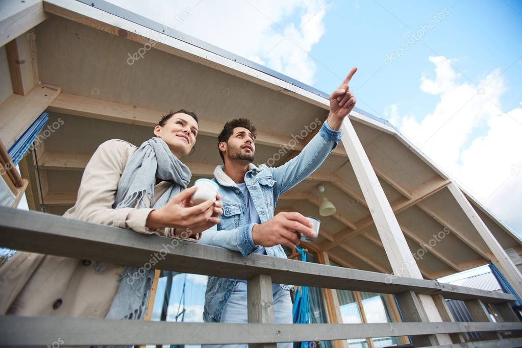 Content handsome young guy with stubble wearing denim jacket leaning on wooden railing and pointing with finger in distance while showing ship to girlfriend, they drinking tea on balcony