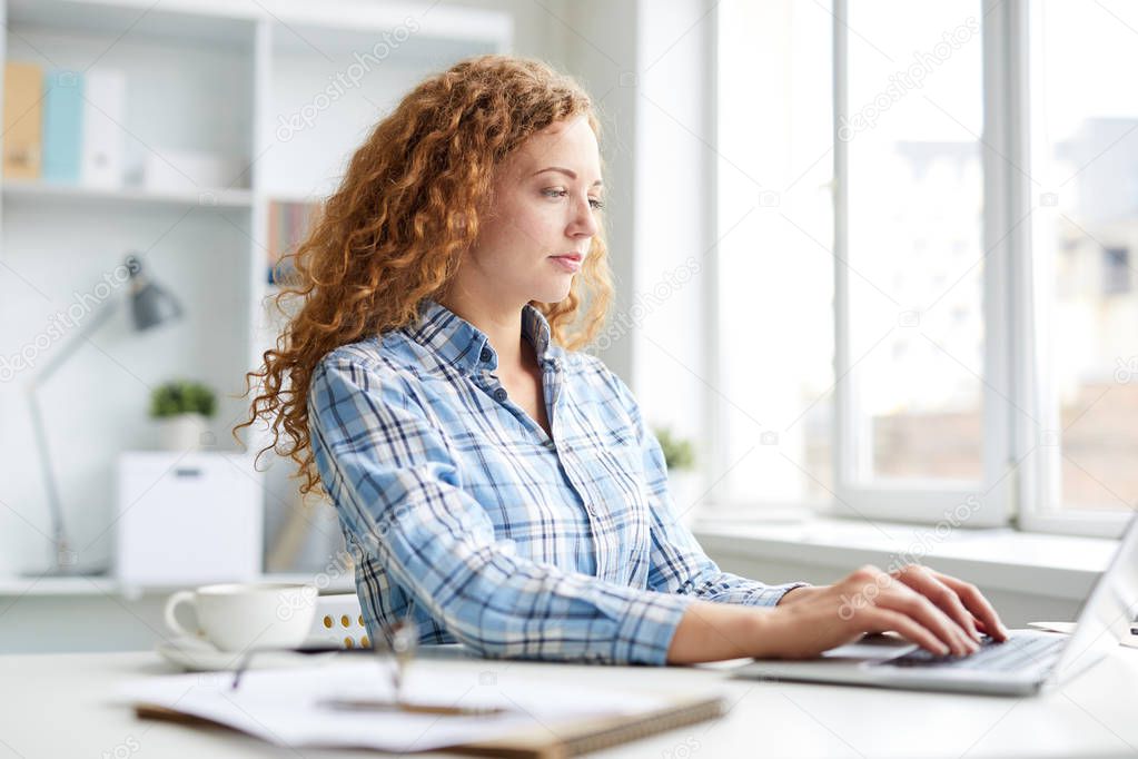 Pretty young serious woman in casualwear sitting by desk in front of laptop and networking