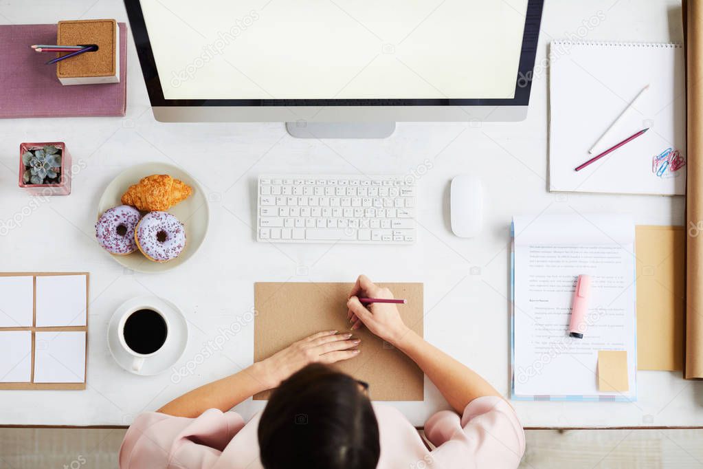 View of busy young manager making notes on paper while sitting in front of computer monitor
