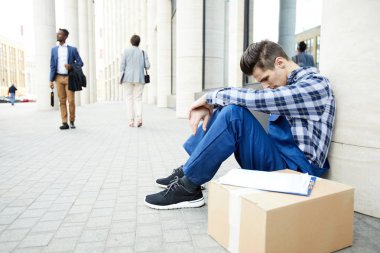 Paketlenmiş kutu ve belge ile modern binalardan birinin duvara oturan iş elbisesi yorgun teslimat adam yakın