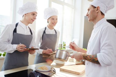 Mutfakta öğrencilere öğretirken soslu tava gösterirken tavsiye veren üniformalı kendinden emin nitelikli yakışıklı şef