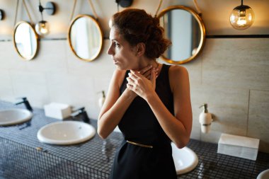 Young brunette woman in elegant black dress touching her throat while standing in toilet clipart