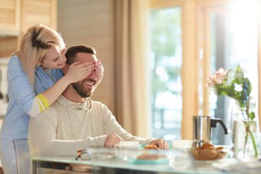 Beyaz sakallı adam mutfak masasında kahvaltı ile oturan ve neşeli gülümseyen karısı arkasında ayakta ve gözlerini kapatırken
