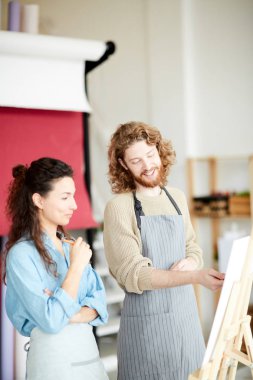 Önlüklü genç adam, ders sırasında tuvale resim çizen arkadaşını işaret ediyor.