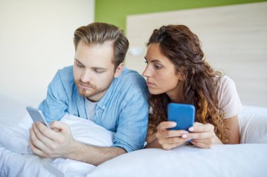 Genç çift yatakta uzanıyor ve akıllı telefonlarıyla arıyor ya da mesajlaşıyor.