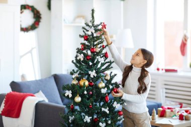 Mutlu kız süslerken Noel ağacının üstüne kırmızı dekoratif yıldız koymak için çalışıyor