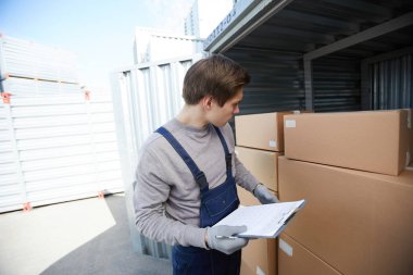 Açık konteynerde dururken kutu sayısını inceleyen ve panoda not lar yapan iş kıyafetinde ciddi odaklı genç erkek depolama yöneticisi