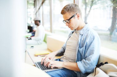 Üniversitede pencerebaşında otururken dizüstü tuş takımı üzerinde yazarak Casual genç adam