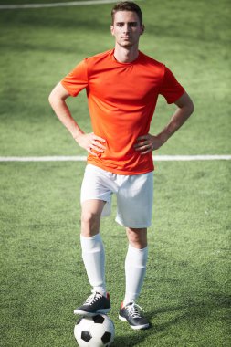 Yeşil sahada ayakta, futbol topu yere bel ve sağ bacak ellerinde tutmak üniformalı genç futbolcu