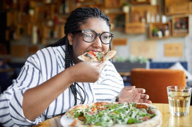 Young woman sitting in cafe and eating appetizing Italian pizza by lunch