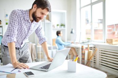 Genç sakallı ofis çalışanı dizüstü bilgisayarla masaya eğilip online verilere bakıyor ya da iş haberlerini izliyor.